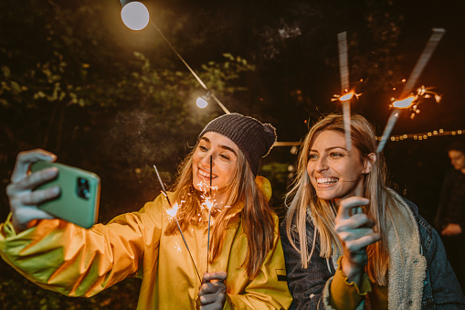 Photo of two women posing and taking pictures while celebrating New Year's with a group of friends; throwing an outdoor party, on a cold evening in the forest; lightning sparklers, dancing, and enjoying each other's company.