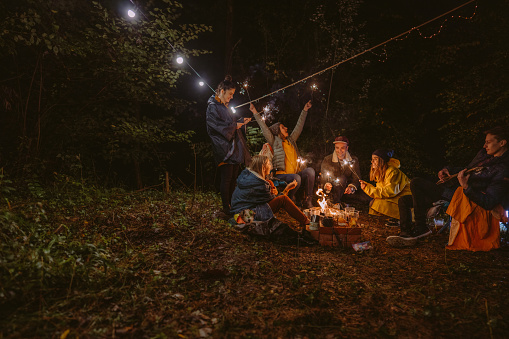 Photo of a group of friends celebrating New Year's by throwing a campfire party in the woods; lightning sparklers while sitting by the campfire, and enjoying each other's company.
