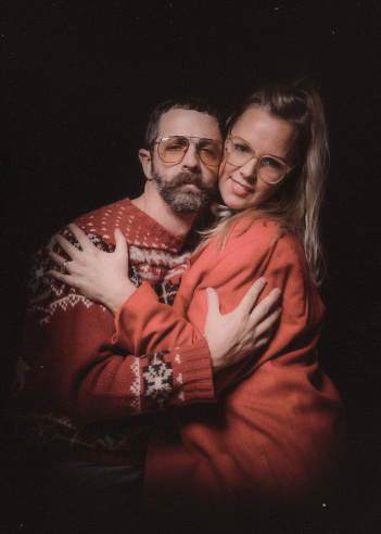 A retro styled image of a couple getting a studio portrait for the holiday season, wearing ugly Christmas sweaters and sitting in an awkward pose.