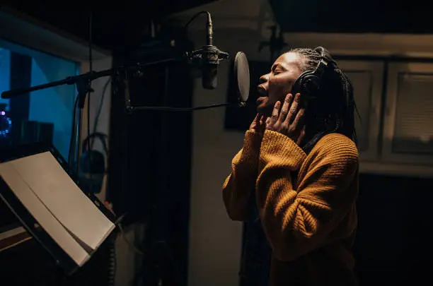 Photo of Black female singer singing into microphone in recording studio