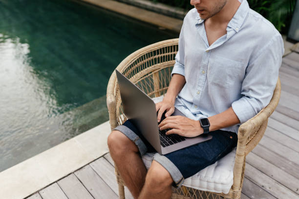 istock Young man using a laptop computer in a garden with a swimming pool. 1284342512