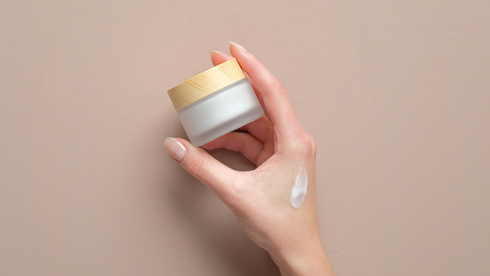Female hand holding jar of cosmetic cream on pastel beige background. Woman applying moisturizer cream