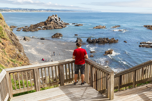 Young man standing on wooden stairs and looking down at Cobble Beach below Yaquina Head lighthouse in Newport, Oregon