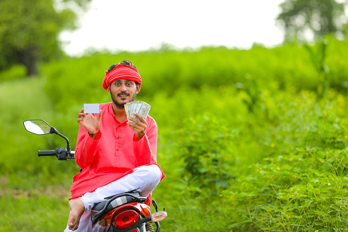 Happy Young indian farmer sitting two wheeler showing money and card at field