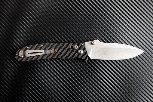 A folding knife with a carbon fiber handle opened up on a carbon fiber background.