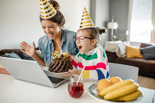 Young mother and daughter are at home, they are wearing party hats and communicating via video call