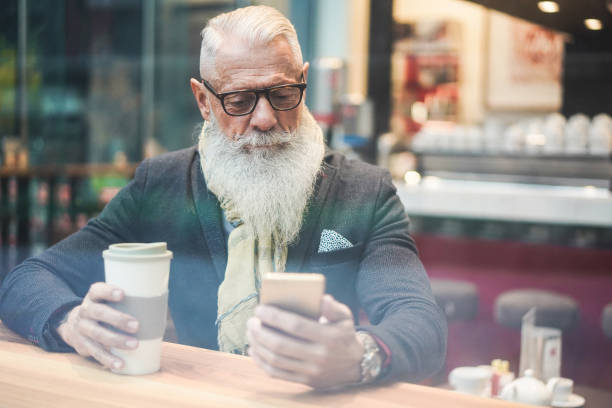 Senior business man using smartphone app while drinking coffee inside cafe bar - Trendy entrepreneur doing breakfast - Job, morning break and tech concept - Window from outside view - Focus on face