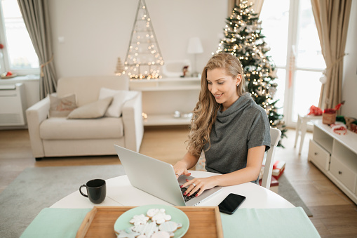Young attractive Caucasian woman casually working from home during Christmas using a laptop, relaxing smiling and enjoying herself
