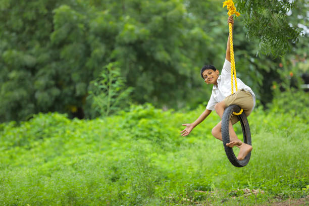 carino bambino indiano che gioca sull'altalena fatta da pneumatico e corda sull'albero sul campo verde - freedom tire swing tire swing foto e immagini stock