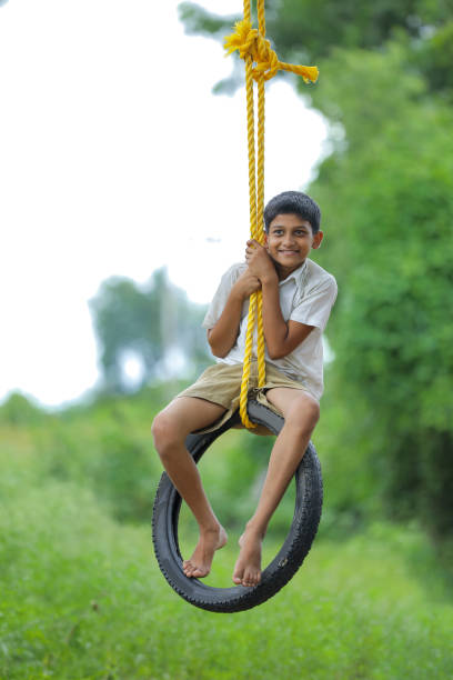 cute indian child playing on swing made by tire and rope on tree at green field - freedom tire swing tire swing imagens e fotografias de stock