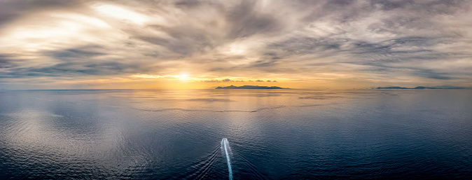 Aerial view of a boat traveling over idyllic, calm ocean towards an lonely island during sunset time with cloudscape in the sky