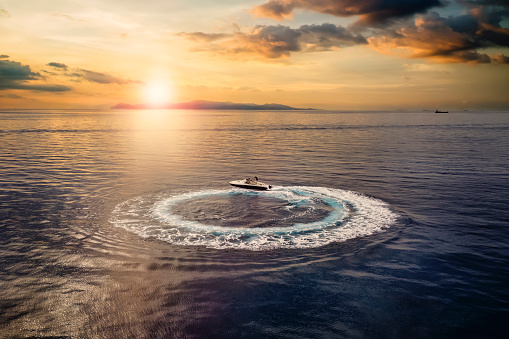 Aerial view of a motorboat forming a circle of waves and bubbles over calm sea during sunset time