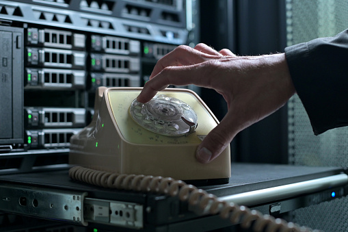 A man puts an old rotary telephone on a shelf against the backdrop of a modern server cabinet and starts dialing a phone number. Clash of eras, progress and technology
