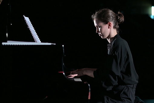 A young woman calmly plays the piano in the spotlight on the stage. Selective lighting, black background
