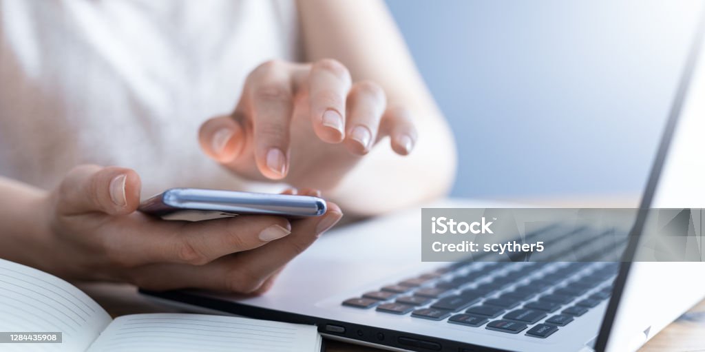 Woman works at home and uses a smart phone and notebook computer. The woman works at home and uses a smart phone and a notebook laptop computer. Silhouette of a busy woman. She holds and using phone. Telephone Stock Photo