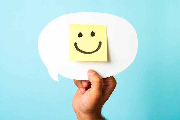 Hand holding white speech bubble with drawing of a happy smiling emoticon on a yellow paper and white background. Hapiness concept. Hand holding white speech bubble with drawing of a happy smiling emoticon on a yellow paper and white background. Hapiness concept. employee engagement stock pictures, royalty-free photos & images