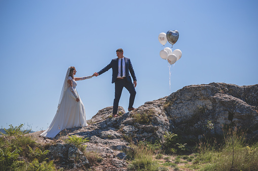 Groom helping bride to climb the rocks. There are balloons waiting.