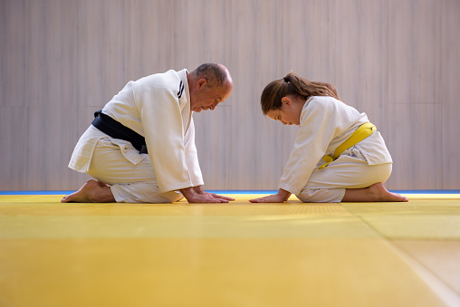 Old Judo sensei with black belt and young female yellow belt student kneeling and bowing to each other showing mutual respect