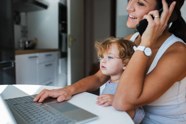 istock Smiling young woman using laptop and talking vis mobile phone when holding a child 1284463829