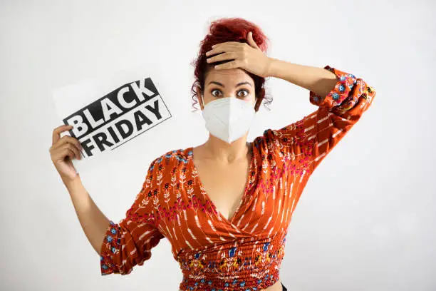 Photo of Girl wearing a mask amazed with wide eyes with one hand on her head and holding a black friday sign with the other hand. Young woman with white skin and curly red hair dressed in colorful patterned clothing