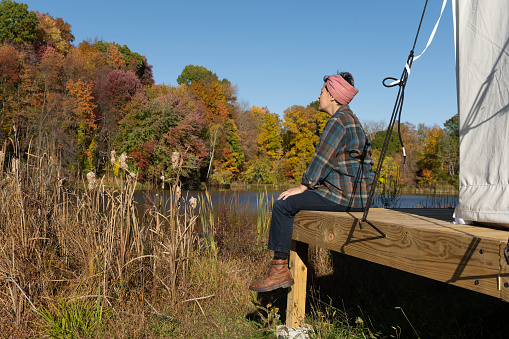 One mixed race non binary female of Persian and Norwegian descent with short hair in a gingham headscarf, a plaid shirt, jeans and boots sits on a simple wooden porch lakeside on a autumn day in the Catskill Mountains while at a glamping campsite on the Stone Ridge Orchard in upstate New York.
