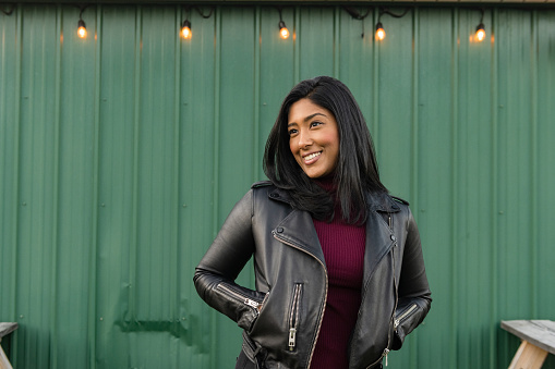 This is a portrait of an Asian American woman of Indian descent in her 30s wearing a black leather jacket and smiling with her hands in her pockets outside a green exterior in the Hudson Valley of upstate New York on an autumn weekend.