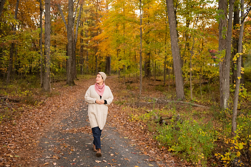A non binary mixed race person wearing a cardigan sweater, scarf and knit hat walks on a country road with autumn foliage in the Hudson Valley of upstate New York.