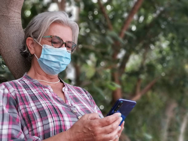 istock Senior woman typing text message on smart phone leaning against a tree trunk. Elderly people and new technology, wearing surgical mask due to coronavirus pandemic 1284511623
