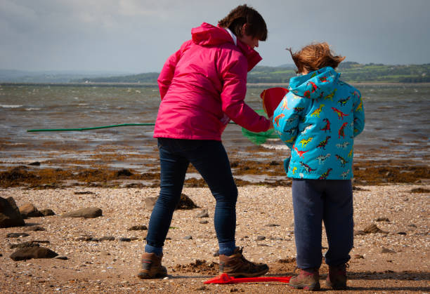 mãe e filha em casacos brilhantes em uma praia irlandesa com uma rede de pesca e balde e pá. - pink pants summer two people - fotografias e filmes do acervo