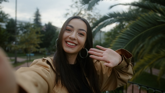 Pretty smiling face woman, holding camera or phone in hand and she is waving to the camera and she pointing out and she makes video call or she is recording video for her fans, technology video call and vlogger concept