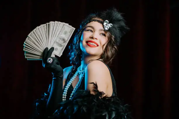 Rich luxurious woman dressed in Gatsby style waves a bundle of dollars banknotes like fan. Vintage, party, retro fashion concept. High quality photo