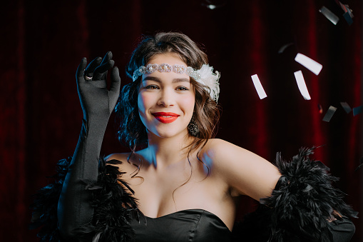 Portrait of retro styled woman dressed in 20th last century dancing under confetti rain on red background. Roaring twenties, retro, party, fashion concept. High quality photo