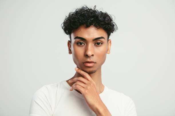 Portrait of an androgynous man on white background Close-up of a handsome androgynous man with his hand on chin. Young guy with feminine facial features looking at camera against white background. queer man facing camera stock pictures, royalty-free photos & images