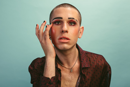 Gender fluid male with makeup looking at camera against blue background. Gay man wearing silk shirt touching his face over blue background.