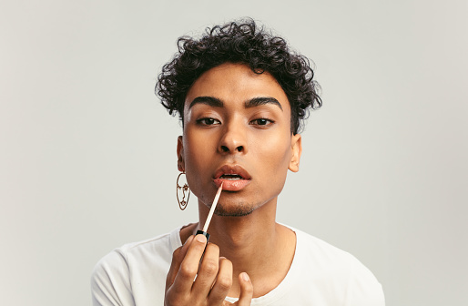 Young man applying lip gloss on his lips and looking in camera like mirror. Gay man doing make up against white background.