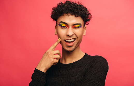 Gay man wearing multicolored shadows on the eyelids winking at camera. Happy transgender male winking an eye against red background.