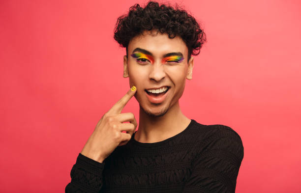 Gay man with rainbow eyeshadow winking at camera Gay man wearing multicolored shadows on the eyelids winking at camera. Happy transgender male winking an eye against red background. man gay stock pictures, royalty-free photos & images