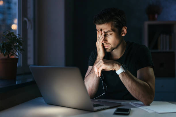 Young overworked man with laptop at night Young businessman has headache while working overtime with laptop at home office late night. Stressed depressed freelancer touching his head, feeling pain in eyes. Insomnia, internet addiction east slavs stock pictures, royalty-free photos & images