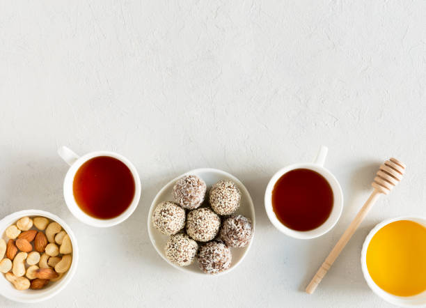 las bolas caseras de energía de caramelo crudo con almendras, anacardos, mantequilla de maní y avellanas en el plato con tazas de té planas yacía sobre fondo gris con espacio de copia. snack orgánico. comida vegetariana. - peanut bowl nut circle fotografías e imágenes de stock