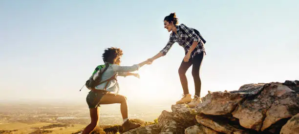 Woman helping her friend to climb the cliff and reach the top of mountain. Friends helping each other during hiking a mountain.