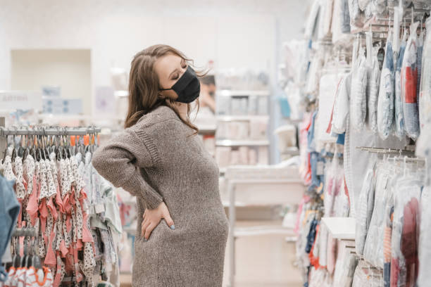 Young beautiful pregnant woman in hygienic mask choosing baby clothes, stroller or pram buggy for newborn. Shopping for expectant mothers and baby. Preventive measures on pandemic lifestyle. stock photo