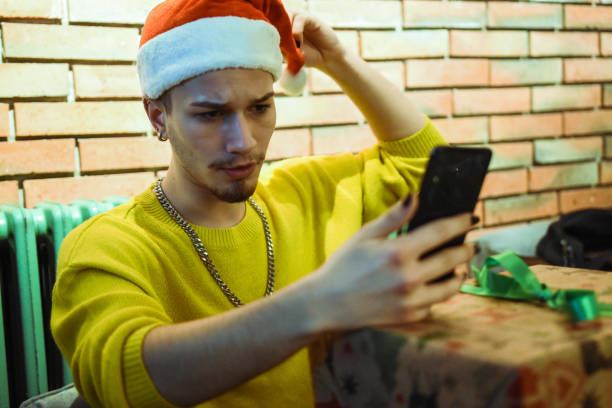 istock Young beautiful queer man with santa hat using phone 1284564330