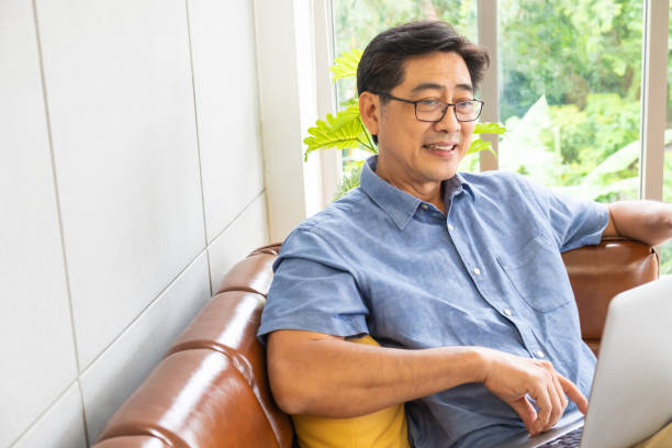 istock Asian senior man sit at sofa couch while using computer laptop for reading news or social network. Active senior with technology concept, Pensioner or retirement life with internet and gadget. 1284571549