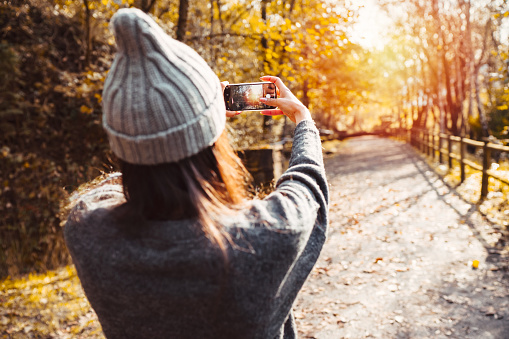 A very beautiful and happy woman in the autumn season on a very bright forest road with orange and yellow tree leaves. The woman is wearing warm clothes and is taking a photo with her smartphone