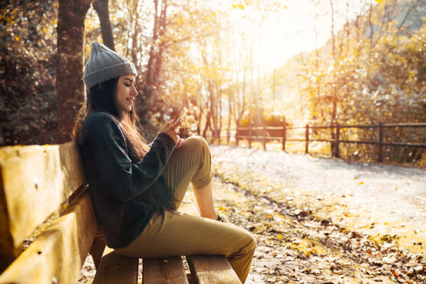 Coat - Garment Very beautiful and very happy woman in the autumn season sitting on the bench of a forest road with the leaves of the orange and yellow trees.The woman is chatting on her mobile phone in warm clothes 21 24 months stock pictures, royalty-free photos & images