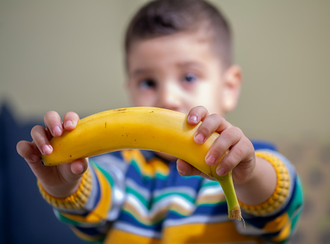 Blurred view of  little boy holding a banana and looking at camera in the house