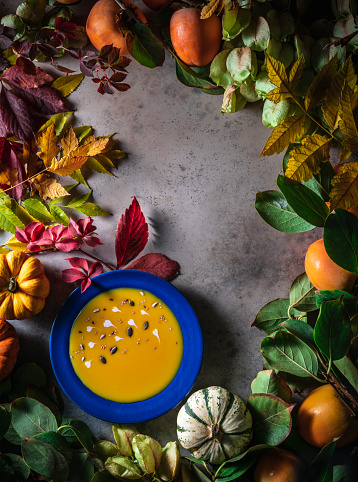Autumn pumpkin soup with raw pumpkins in a persimmon harvest background with autumn leaves in a blue plate