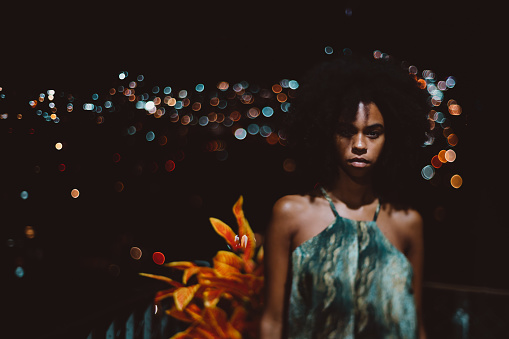 A shot in a dark key of a young ravishing Brazilian female in a simple sundress standing on her house balcony at warm night, with a cityscape behind her, totally blurred into bokeh bulbs