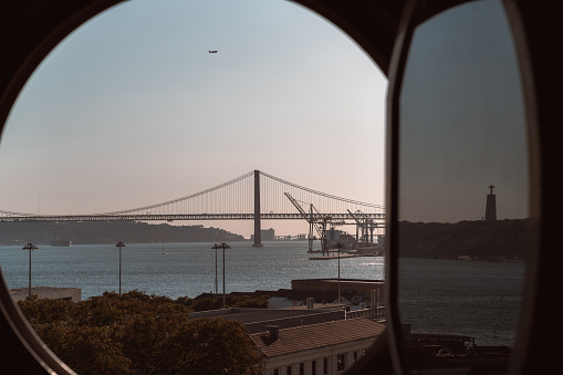View through opened round illuminator window of an old historic building of an evening Lisbon cityscape with a bridge over the Tejo river in the distance, water, descending airplane
