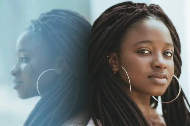 A close-up portrait of a cute young black woman with dreadlocks and huge earrings, piercing in her nose, she is leaning against a glass wall outdoors which fully reflects her and looking at the camera